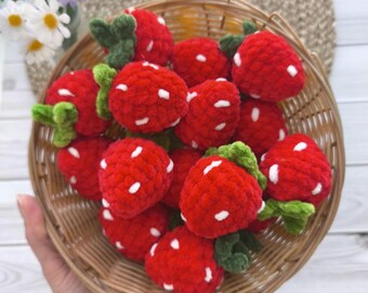 Puede incluir: Una cesta de mimbre llena de fresas rojas hechas a ganchillo con hojas verdes y puntos blancos. La cesta está sostenida por una persona. Las margaritas y las flores moradas están en el fondo.