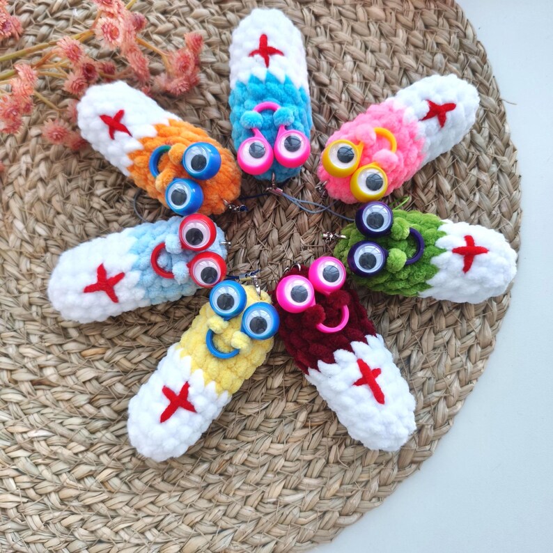 May include: A collection of colorful, handmade crochet keychains. Each keychain features a pill-shaped design with a smiling face, googly eyes, and a red cross. The keychains are in various colors, including orange, blue, pink, green, yellow, and burgundy.