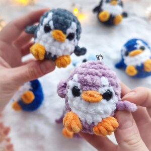 May include: A collection of handmade crochet penguin ornaments in various colors. The penguins have white bellies, black eyes, and orange feet and beaks. One penguin is held in a person's hands. Other penguins are blue, black, and purple.
