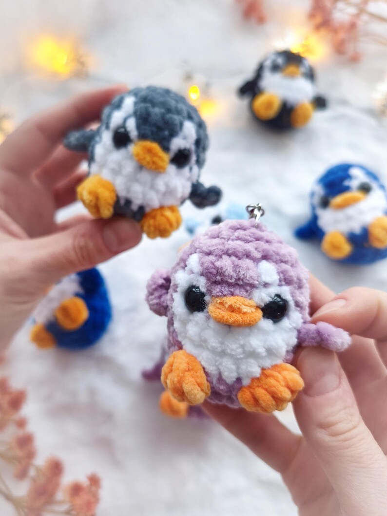 May include: A collection of handmade crochet penguin ornaments in various colors. The penguins have white bellies, black eyes, and orange feet and beaks. One penguin is held in a person's hands. Other penguins are blue, black, and purple.
