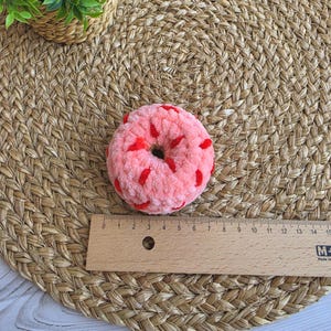 Puede incluir: Un juguete de rosquilla rosa de ganchillo suave y afelpado con chispas de hilo rojo. Una regla de madera est&aacute; colocada directamente debajo de la rosquilla, mostrando que mide aproximadamente 9 cm de di&aacute;metro. Tiene forma circular.