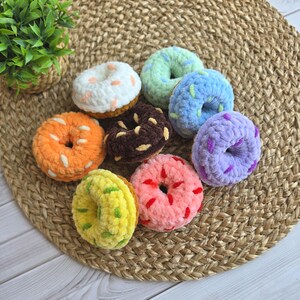 Puede incluir: Una colecci&oacute;n de ocho rosquillas de peluche tejidas a ganchillo, de forma redonda y colores variados que incluyen blanco, naranja, marr&oacute;n oscuro, verde claro, azul, morado, rosa y amarillo, con virutas de lana en contraste sobre la superficie.