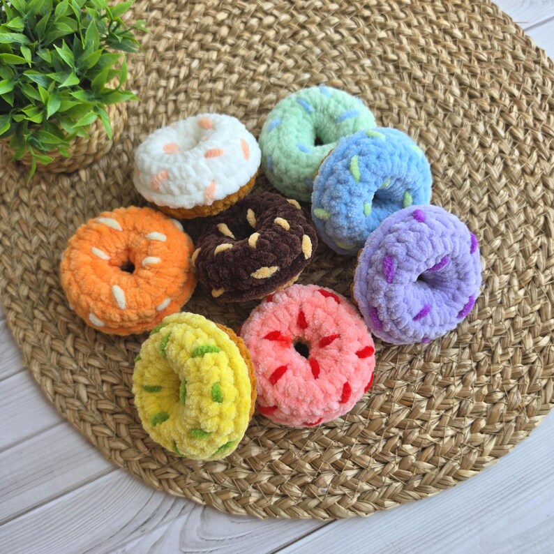 Puede incluir: Una colecci&oacute;n de ocho rosquillas de peluche tejidas a ganchillo, de forma redonda y colores variados que incluyen blanco, naranja, marr&oacute;n oscuro, verde claro, azul, morado, rosa y amarillo, con virutas de lana en contraste sobre la superficie.