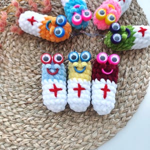 May include: A collection of colorful, handmade crochet pill-shaped keychains. Each keychain features a smiling face, googly eyes, and a red cross design. The keychains are in various colors, including blue, yellow, and red.