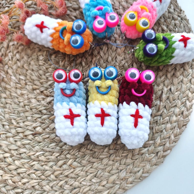 May include: A collection of colorful, handmade crochet pill-shaped keychains. Each keychain features a smiling face, googly eyes, and a red cross design. The keychains are in various colors, including blue, yellow, and red.