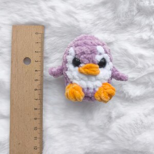 May include: A handmade, plush penguin toy in shades of purple, white, and orange. The penguin has a soft, textured body, with a white belly and head, and orange feet and beak. The toy is approximately 4 inches tall.