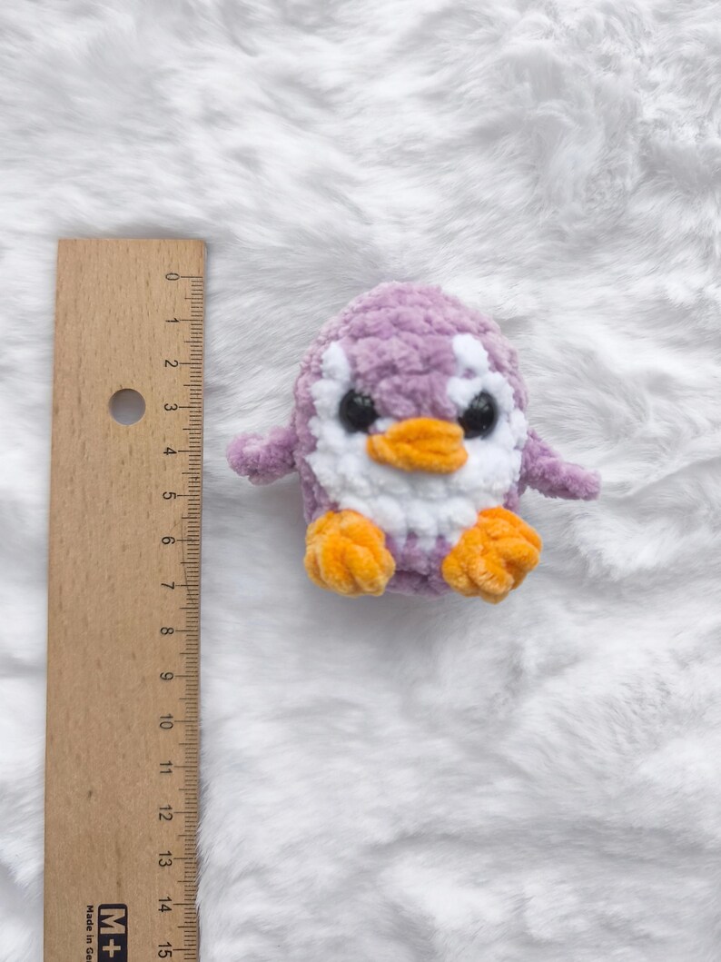 May include: A handmade, plush penguin toy in shades of purple, white, and orange. The penguin has a soft, textured body, with a white belly and head, and orange feet and beak. The toy is approximately 4 inches tall.