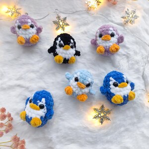 May include: A collection of seven crocheted penguin ornaments in various colors, including purple, blue, and black. Each penguin has a white belly, yellow feet, and small black eyes. The penguins are arranged on a white, fluffy surface with snowflake lights.