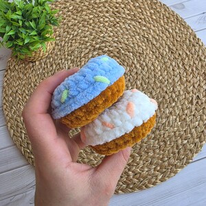 Puede incluir: Dos art&iacute;culos de ganchillo con forma de cupcake, bases marrones y partes superiores texturizadas. Un cupcake tiene la parte superior azul claro con peque&ntilde;os detalles verdes y el otro blanca con detalles color melocot&oacute;n, hechos de lana suave.
