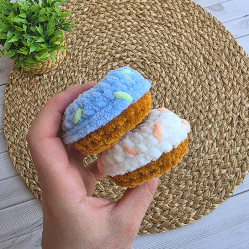 Puede incluir: Dos art&iacute;culos de ganchillo con forma de cupcake, bases marrones y partes superiores texturizadas. Un cupcake tiene la parte superior azul claro con peque&ntilde;os detalles verdes y el otro blanca con detalles color melocot&oacute;n, hechos de lana suave.