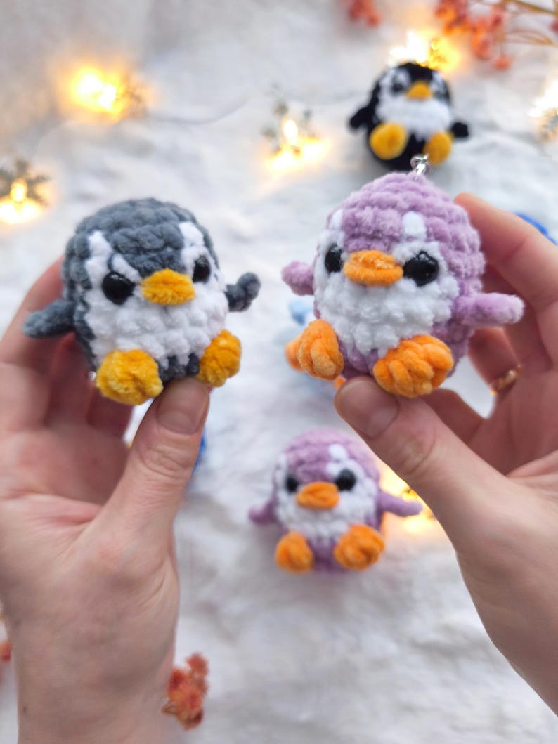 May include: Handmade crochet penguin ornaments in various colors: gray, purple, and black. Each penguin has a white belly, orange feet and beak, and black eyes. These small, plush penguins are perfect for holiday decorations or as a cute gift.