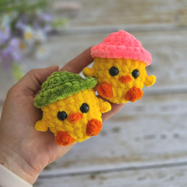 May include: Two small, yellow, crocheted plush chicks with textured yarn. One chick wears a green hat, and the other wears a bright pink hat. Both have black plastic eyes, orange beaks, and small orange feet, crafted from soft, fuzzy material.