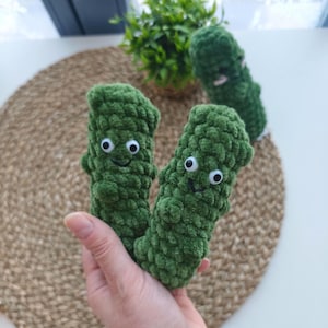 Könnte beinhalten: Handgemachte grüne Häkelgurkenspielzeuge mit Wackelaugen und lächelnden Gesichtern. Die Plüschgurken sind aus einem weichen, strukturierten Garn gefertigt. Das Bild zeigt zwei Gurken in einer Hand, mit einer dritten Gurke und einer Pflanze im Hintergrund.