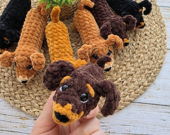 May include: A collection of handmade crochet dachshund dog toys in various shades of brown and black. The toys have floppy ears, small black eyes, and a cute, elongated body. The toys are arranged on a woven mat.