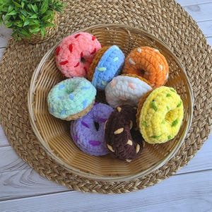 Puede incluir: Una colecci&oacute;n de ocho juguetes de rosquillas de ganchillo de felpa en varios colores, incluyendo rosa, azul, naranja, blanco, verde menta, morado, marr&oacute;n y amarillo, cada uno con chispas de hilo contrastantes, dispuestos en una cesta de mimbre.