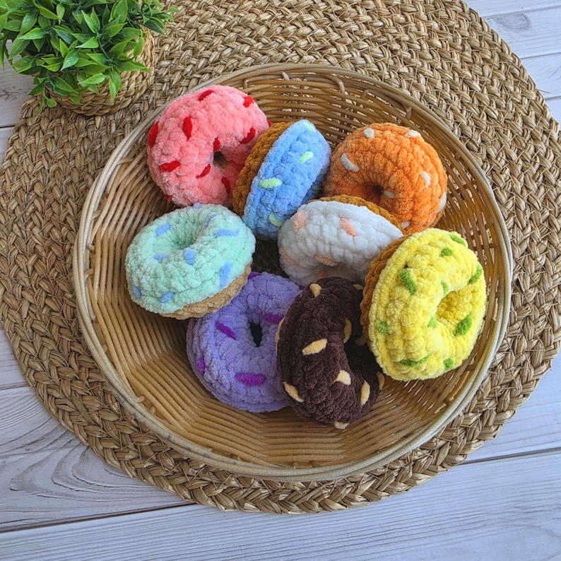 Puede incluir: Una colecci&oacute;n de ocho juguetes de rosquillas de ganchillo de felpa en varios colores, incluyendo rosa, azul, naranja, blanco, verde menta, morado, marr&oacute;n y amarillo, cada uno con chispas de hilo contrastantes, dispuestos en una cesta de mimbre.
