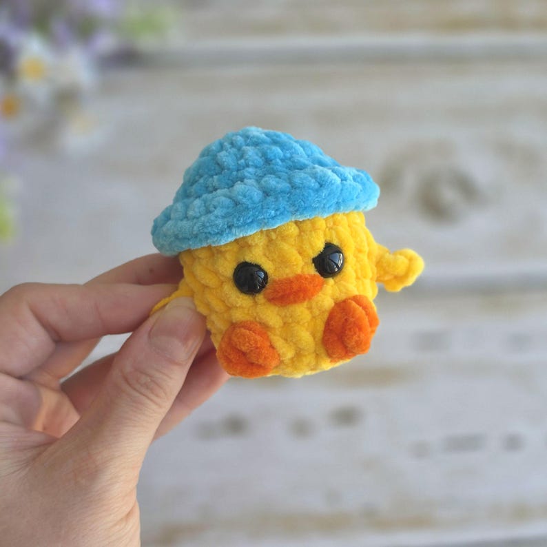 May include: A small, handmade crochet duckling toy crafted from soft yellow yarn. The duckling features a bright blue crocheted hat, two round black plastic eyes, a small orange beak, and two orange feet. The texture is fuzzy and plush throughout.