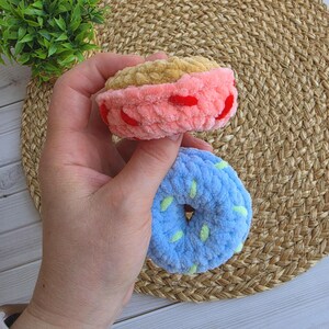 Puede incluir: Dos juguetes de rosquilla tejidos a ganchillo con hilo de felpa suave. La rosquilla superior es de color tostado con glaseado rosa y chispas rojas. La inferior es azul claro con chispas verde p&aacute;lido. Ambos tienen una textura gruesa y afelpada.