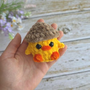 May include: A small, handmade crochet plush toy shaped like a yellow chick wearing a brown textured acorn cap. The chick features two round black plastic eyes, a small orange beak, and two orange feet, all crafted from soft, fuzzy yarn material.
