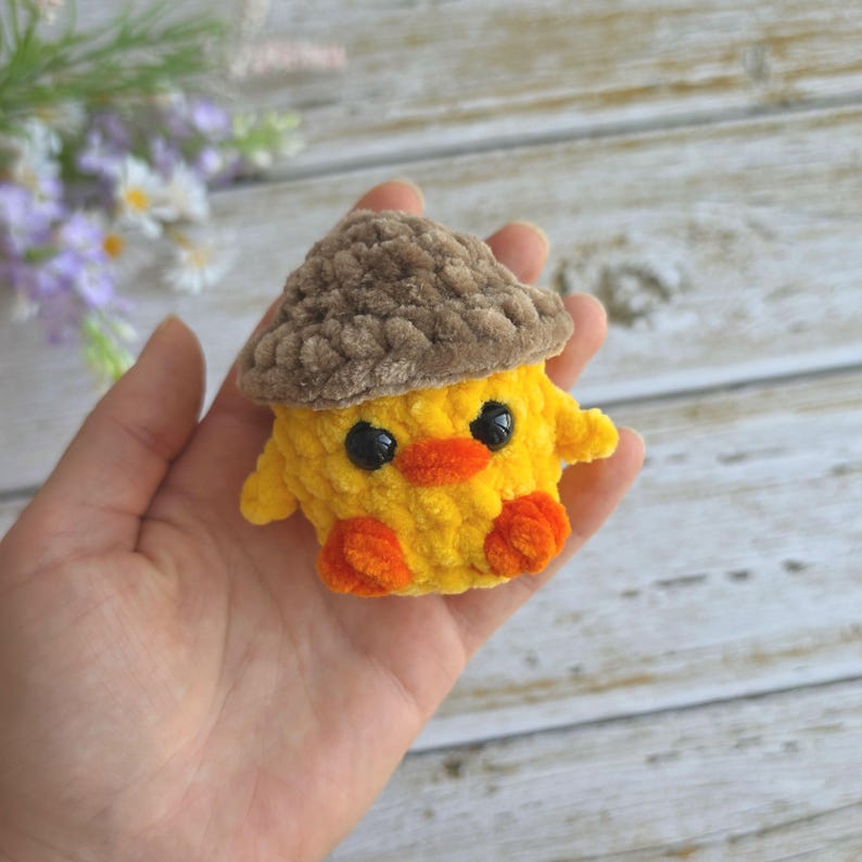 May include: A small, handmade crochet plush toy shaped like a yellow chick wearing a brown textured acorn cap. The chick features two round black plastic eyes, a small orange beak, and two orange feet, all crafted from soft, fuzzy yarn material.
