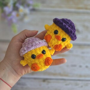 May include: Two small, yellow, crocheted ducklings made from soft, plush yarn. Each duckling features bright orange beaks and feet, black bead eyes, and a textured hat. One duckling wears a light lavender hat, while the other wears a dark purple hat.