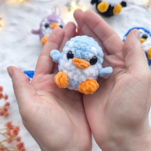 May include: A small, handmade crochet penguin in light blue, white, and orange. The penguin is held in open hands, with other crochet penguins in the background. The penguin has black eyes and orange feet and beak.