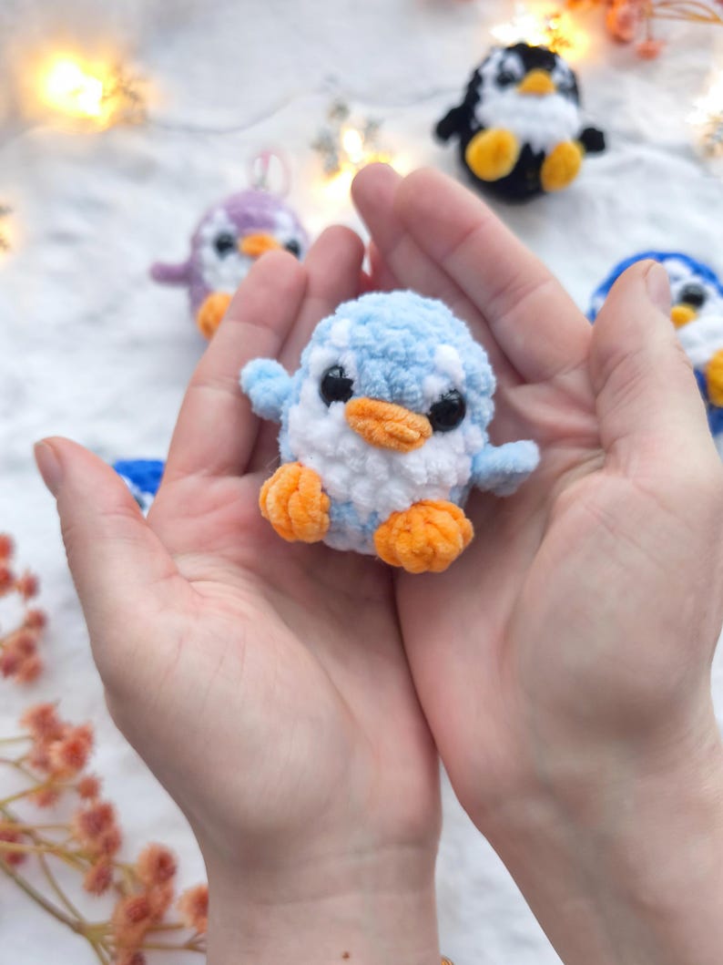 May include: A small, handmade crochet penguin in light blue, white, and orange. The penguin is held in open hands, with other crochet penguins in the background. The penguin has black eyes and orange feet and beak.