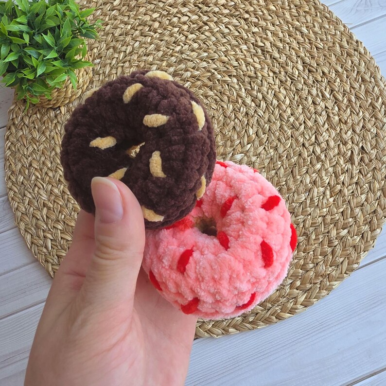 Puede incluir: Dos juguetes de rosquilla tejidos a ganchillo con hilo de felpa suave. Una rosquilla es de color marr&oacute;n oscuro con chispas de color canela claro, y la otra es de color rosa claro con chispas rojas brillantes. Ambas tienen forma circular.