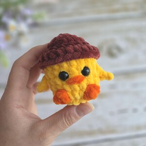 May include: A small crocheted plush toy shaped like a yellow chick wearing a dark brown acorn cap. The chick features two round black plastic eyes, a small orange beak, and two orange feet. The texture is soft and fuzzy with visible yarn loops.