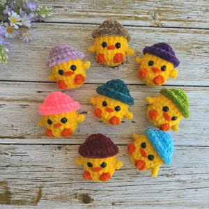 May include: Eight small, round, yellow crocheted chicks, each wearing a different colored hat. The hats are brown, purple, dark green, light green, blue, maroon, pink, and lavender. Each chick has black bead eyes and orange feet.