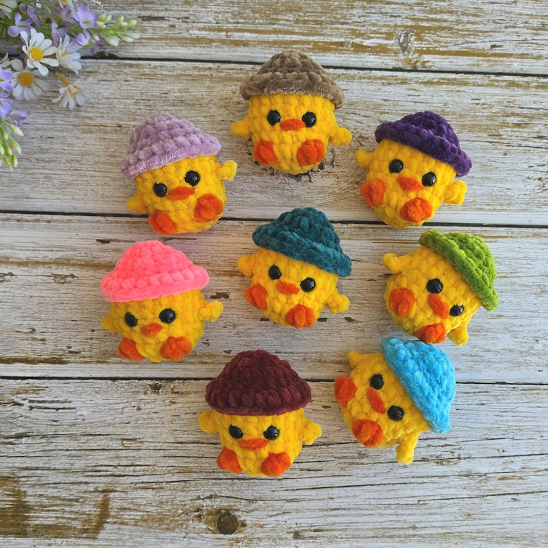 May include: Eight small, round, yellow crocheted chicks, each wearing a different colored hat. The hats are brown, purple, dark green, light green, blue, maroon, pink, and lavender. Each chick has black bead eyes and orange feet.