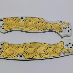 May include: Two metallic knife handle scales featuring a silver-toned border and a central gold-colored inlay with a repeating embossed scale or leaf pattern. Each piece includes pre-drilled circular holes for assembly and mounting hardware.