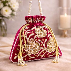 Könnte beinhalten: Eine bordeauxfarbene Samt-Beuteltasche mit goldener Blumenstickerei und einem perlenbesetzten Griff. Die Tasche hat einen Kordelzugverschluss mit goldener Kordel und Perlenquasten. Die Tasche ist mit weißen Perlen an den Rändern verziert.