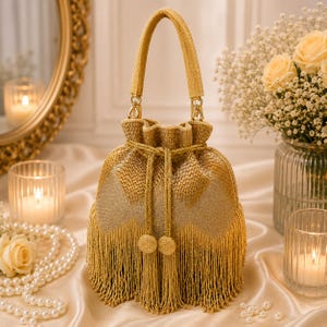 May include: A gold-coloured drawstring handbag with a woven texture and long fringe. The bag has a matching handle and gold-coloured hardware. The bag is decorated with two tassel-like embellishments. Set against a soft, neutral background.