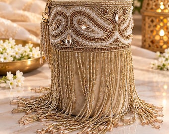 May include: A cylindrical handbag featuring a beige base adorned with gold and white beadwork in a paisley pattern. The upper section is embellished with clear crystals, while the lower portion is covered in long, dangling gold beaded fringe.
