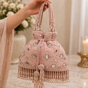 Puede incluir: Un bolso de mano rosa rubor con cuentas, cierre de cordón y detalles de perlas. El bolso presenta un patrón decorativo de cuadrados espejados y adornos redondos. Las asas también están adornadas con cuentas y perlas, y la parte inferior tiene una borla de perlas.