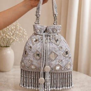 May include: A silver beaded drawstring bag with a decorative pattern of mirrored squares and round embellishments. The bag has a beaded fringe and beaded handles. The bag is held up by a hand.
