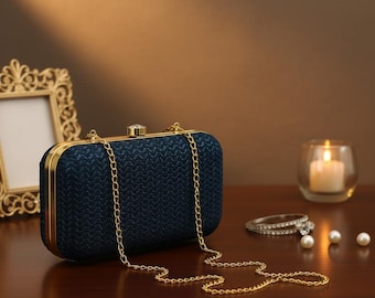 May include: A navy blue rectangular clutch with a gold chain strap. The clutch has a textured woven pattern and gold trim. A gold picture frame, a candle in a glass holder, and jewelry are in the background.