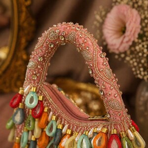 May include: A pink velvet handbag featuring a top handle embellished with intricate gold beadwork and clear crystals. The bag is adorned with a fringe of dangling, teardrop-shaped charms in vibrant red, orange, green, and blue colors.