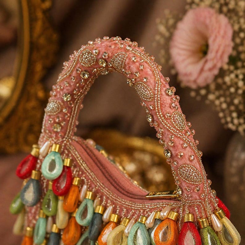 May include: A pink velvet handbag featuring a top handle embellished with intricate gold beadwork and clear crystals. The bag is adorned with a fringe of dangling, teardrop-shaped charms in vibrant red, orange, green, and blue colors.