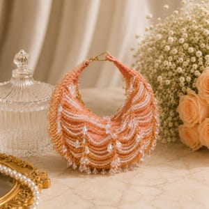 May include: A peach-colored, crescent-shaped handbag with a gold chain handle. The bag is embellished with rows of white and gold beads, and dangling crystal accents. A decorative glass jar and a bouquet of roses are in the background.