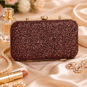 Puede incluir: Un clutch rectangular burdeos brillante con herrajes dorados. El clutch está rodeado de accesorios, incluyendo un labial, un collar de perlas y aretes, todo sobre una superficie satinada. Un frasco de perfume y flores están en el fondo.