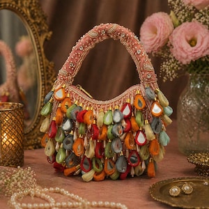 May include: A pink handbag featuring a top handle adorned with intricate beadwork and crystals. The exterior is covered in numerous dangling, teardrop-shaped fabric charms in shades of red, orange, green, and gray, interspersed with small mirrors.