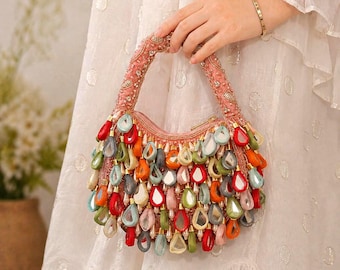 May include: A crescent-shaped handbag in a soft pink hue, featuring an embellished handle and a dense arrangement of dangling, teardrop-shaped charms in vibrant shades of red, orange, green, blue, and white, each with a reflective center.