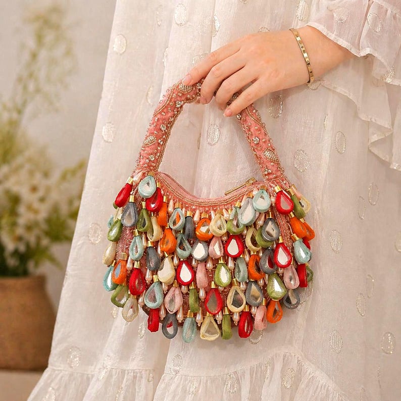 May include: A crescent-shaped handbag in a soft pink hue, featuring an embellished handle and a dense arrangement of dangling, teardrop-shaped charms in vibrant shades of red, orange, green, blue, and white, each with a reflective center.