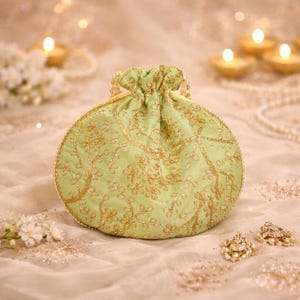 Puede incluir: Un pequeño bolso redondo verde claro con cordón y bordado floral dorado. El bolso tiene un ribete de cuentas doradas y un cierre de cordón dorado. El fondo incluye velas y elementos decorativos.
