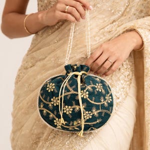 May include: A dark teal velvet pouch bag with gold floral embroidery and a pearl beaded handle. The bag has a drawstring closure and is held against a cream-coloured, embroidered fabric. The bag's design is ornate and elegant.