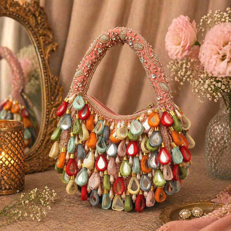 May include: A pink handbag featuring a top handle adorned with clear crystals and a body covered in dangling, teardrop-shaped charms in various colors including orange, green, red, and light blue, each with a central reflective mirror inlay.
