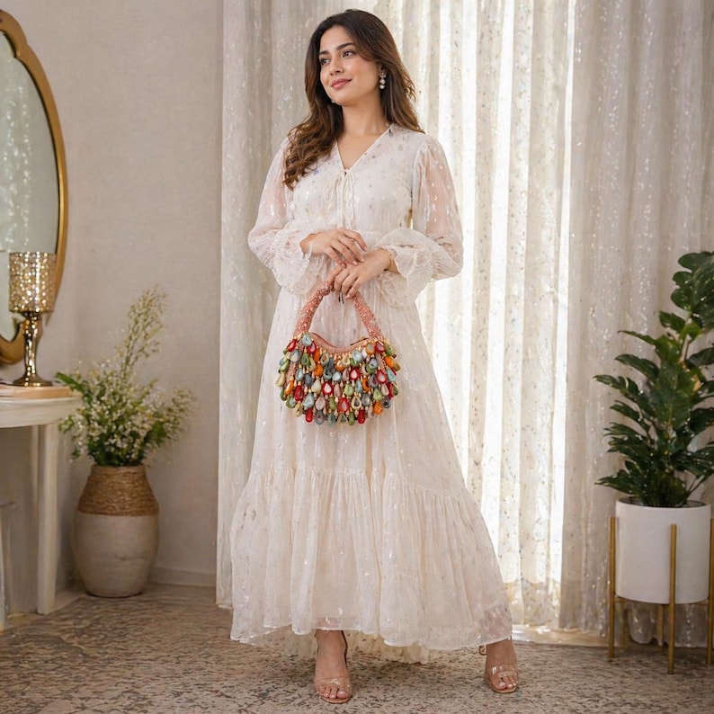 May include: A long-sleeved, floor-length cream dress with a subtle metallic pattern, featuring a tiered skirt and a V-neckline. The dress is paired with a crescent-shaped handbag adorned with numerous colorful, teardrop-shaped embellishments.