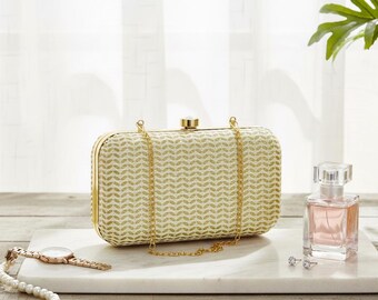 May include: A rectangular clutch with a gold frame and chain strap. The clutch has a cream-colored background with a gold chevron pattern. A gold watch and a perfume bottle are also in the image.